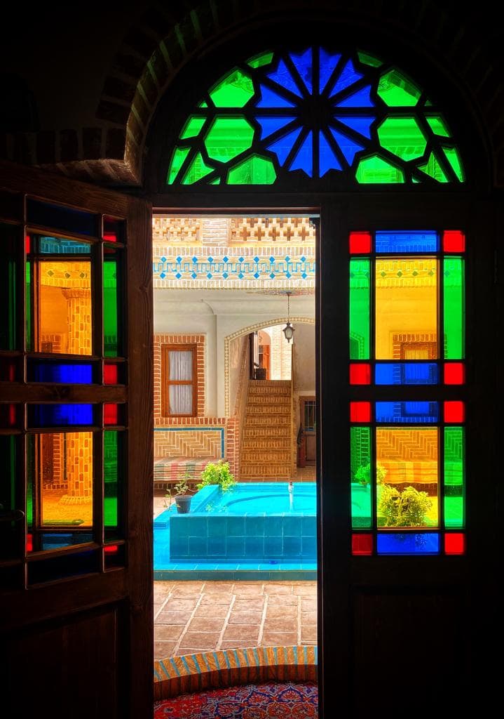 نمای حیاط و حوض فیروزه‌ای از پشت در ارسی رنگی